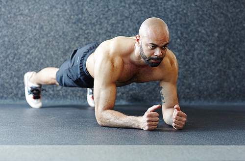 Motivational portrait of shirtless muscular man holding plank  against grey background, straining with effort and looking determined during intense endurance workout in gym