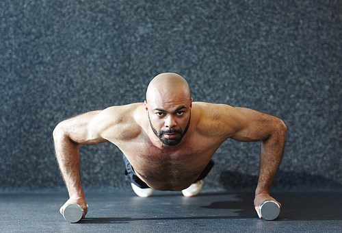 Motivational portrait of shirtless muscular man performing push-ups  on dumbbells and  ,straining with effort during intense endurance workout in gym