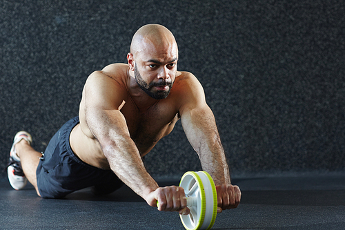Portrait of muscular shirtless man exercising with hand roller on floor against grey background in gym