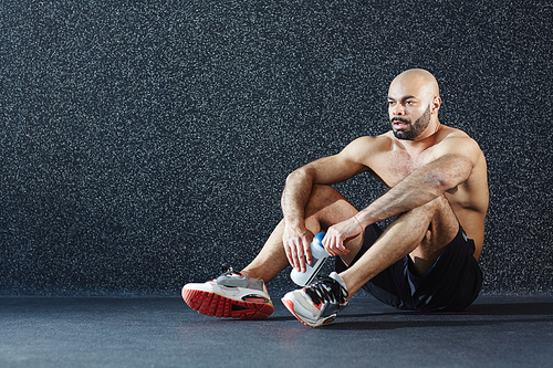 Portrait of handsome muscular athlete sitting on floor, resting after intense workout in gym