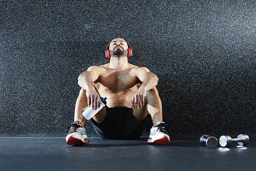 Young athlete leaning against wall while sitting on the floor and listening to music in headphones