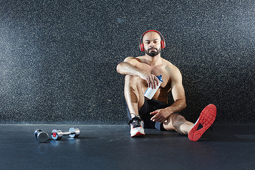 Muscular man with headphones and bottle of water sitting on the floor