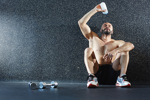 Portrait of shirtless muscular man sitting on floor pouring water on face refreshing after  intense strength workout in gym
