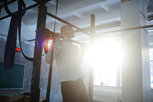 Portrait of bald bearded man performing pull ups on bar during workout in gym lit by sunlight with lens flare