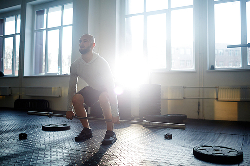 Portrait of strong bearded man lifting barbell during crossfit workout in gym lit by sunlight