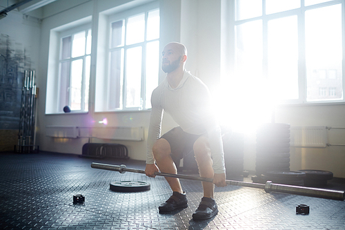 Motivational portrait of strong bearded man lifting barbell during workout in gym lit by sunlight