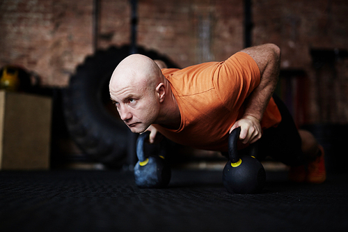 Bald sporty man standing in plank position with help of kettlebells and looking away with concentration, full-length portrait