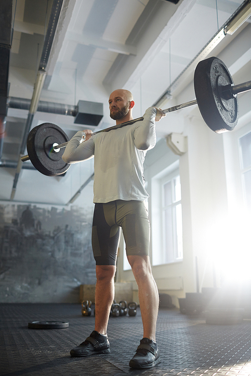 Low angle portrait of modern strongman weightlifting with heavy barbell performing shoulder press during powerlifting workout in sunlit gym