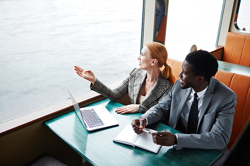 Businesswoman pointing at waterside behind window while talking to colleague during business travel on steamer