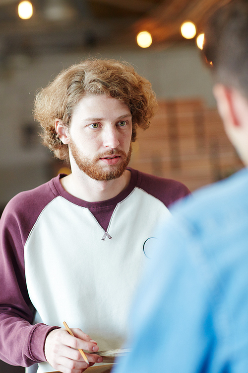 Waist-up portrait of bearded red-haired manager having project discussion with his colleague and taking necessary notes, blurred background