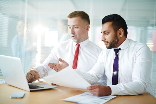 Two businessmen sitting in front of laptop and looking through online financial statistics in office