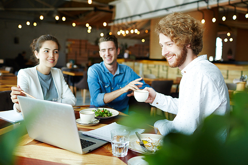 Young man with cup of tea looking at data on laptop display being pointed at by one of colleagues at meeting