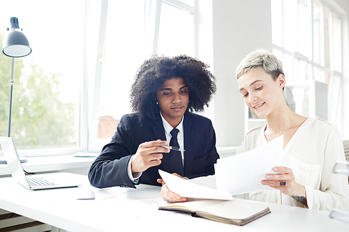 Hard-working group of managers sitting at desk and analyzing results of accomplished work, interior of spacious open plan office on background