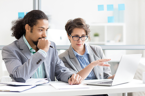 Confident employee making presentation and pointing at data on laptop display while explaining it to colleague