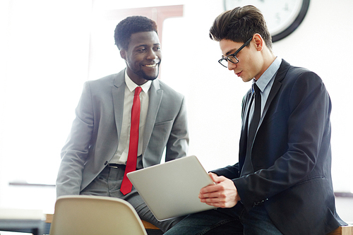 Multi-ethnic group of coworkers wearing suits discussing results of accomplished work with help of laptop while having working meeting at spacious boardroom