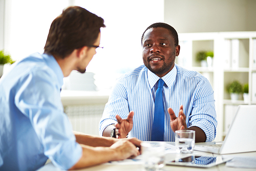 Multiethnic group of businesspeople brainstorming in spacious meeting room, table with laptop, digital tablet, documents and glasses of water on foreground