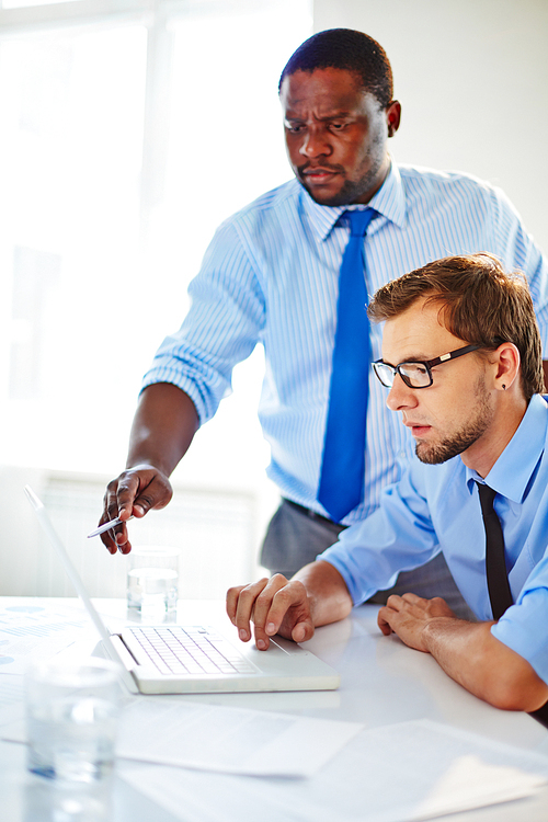 Dissatisfied African American boss pointing at mistake displayed on screen, bearded employee in eyeglasses sitting in front of laptop and making corrections