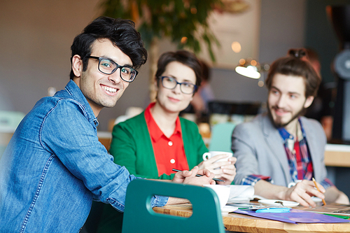 Portrait of three creative people dressed in business casual working at table during meeting, focus on young smiling man wearing glasses