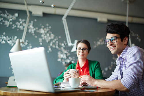 Portrait of two young creative colleagues man and woman both wearing casual clothes and glasses, working at meeting in cafe, smiling  and looking at laptop screen