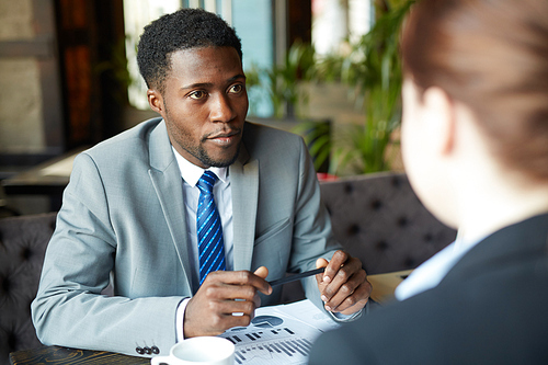 Two business people meeting in modern cafe: African-American man wearing business suit looking at his partner listening intently