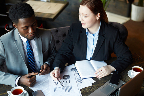 Portrait of two business people meeting in modern cafe: Young professional woman pointing to statistics graphs while explaining data to African ?American colleague