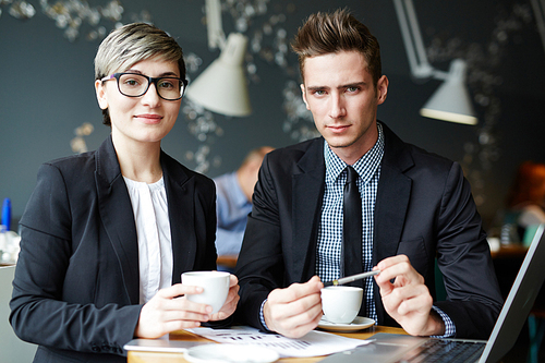 Male and female manages posing for photography while drinking coffee in small cafe and working on their joint project, waist-up portrait