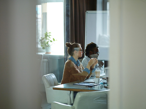 Side view of modern young people in casual clothes sitting at meeting table in conference room listening intently to speaker, shot from behind glass door