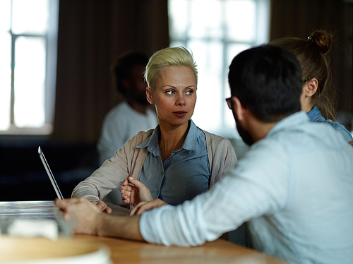 Portrait of several modern business people dressed in casual clothes sitting together at table in dim room, having discussion meeting and talking to each other sharing ideas, focus on blond woman wearing creative haircut