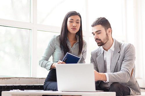 Productive teamwork in spacious office: concentrated Asian manager with notebook and pen in hands looking at screen of laptop with interest while her bearded colleague showing his work results