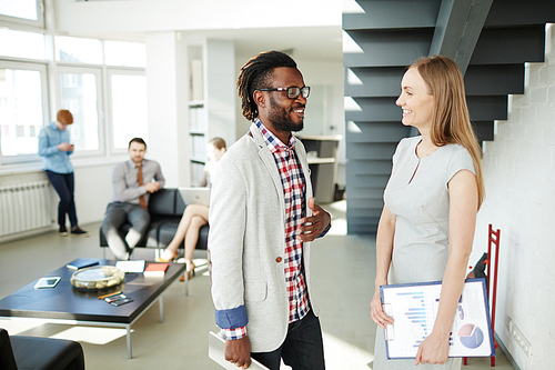 Having break after productive business meeting: Afro-American employee and his pretty female colleague chatting on foreground while other workers seen on background