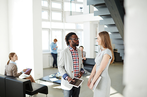 Pretty businesswoman with laptop standing next to Afro-American colleague with digital tablet while having informal meeting in office lobby