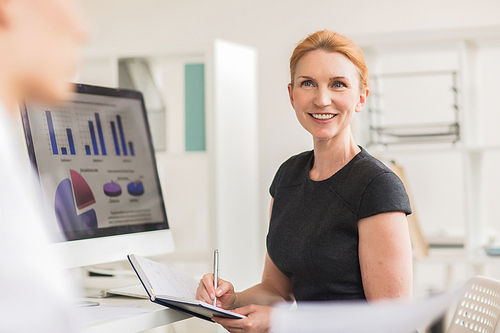 Cheerful middle-aged manager looking at her colleague with charming smile and taking notes while he presenting ideas concerning their joint project, interior of open plan office on background