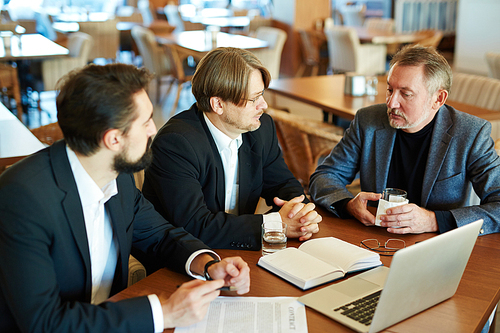 Three confident business partners in formalwear discussing new contract terms before signing it while sitting in spacious restaurant, waist-up portrait