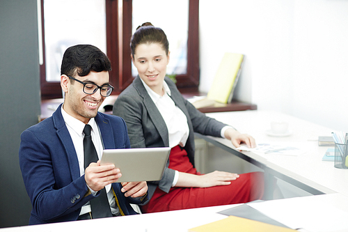 Handsome bearded designer with digital tablet in hands showing funny video to his pretty coworker while taking short break from work, interior of modern open plan office on background