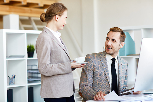 Pretty young woman with cup of fragrant coffee chatting animatedly with her handsome bearded colleague, interior of modern office on background