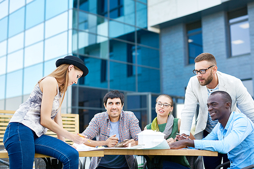 Multi-ethnic group of carefree young people working together at outdoor table with house model on it collaborating on college project by modern glass building