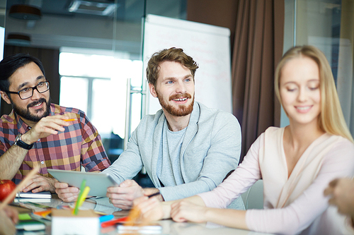 International group of modern, casually dressed people, two young men and woman, sitting at table during business meeting with colleagues, discussing project enthusiastically and sharing ideas