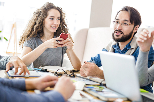 Young businesswoman with smartphone listening to her colleagues conversation at meeting