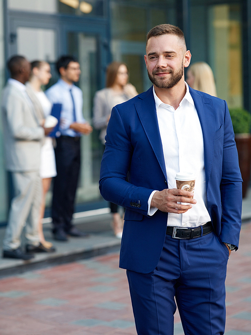 Portrait of young handsome man wearing stylish blue suit smiling and drinking coffee from paper cup outside office building, group of people in background