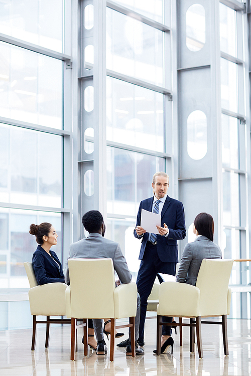 Group of  business people sitting in circle during meeting, man standing up giving presentation in glass hall of modern office building