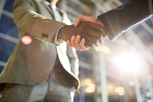 Low angle closeup shot of two business partners in handshake: unrecognizable African -American businessman shaking hands with Caucasian colleague in hall of modern glass office building at night time