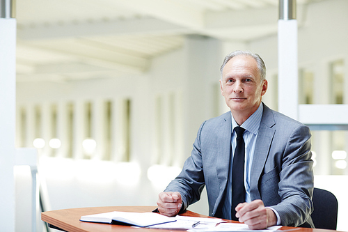 Mature boss in formalwear sitting by his workplace in office and planning work