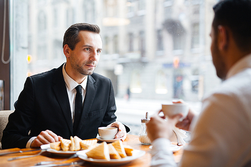 Portrait in warm lighting of two businessmen in formalwear discussing important matters in cafe over a light meal, focus on intent dark-haired man listening to his colleague