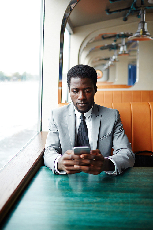 Posh passenger of steamship sitting by window and texting in smartphone