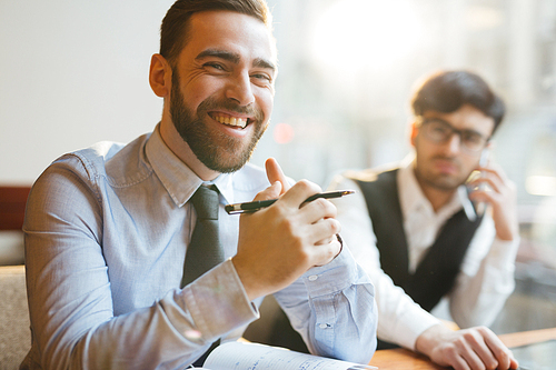 Portrait of young attractive company executive sitting at table with clasped hands, holding pen and smiling at camera cheerfully, sitting next to his pensive assistant.