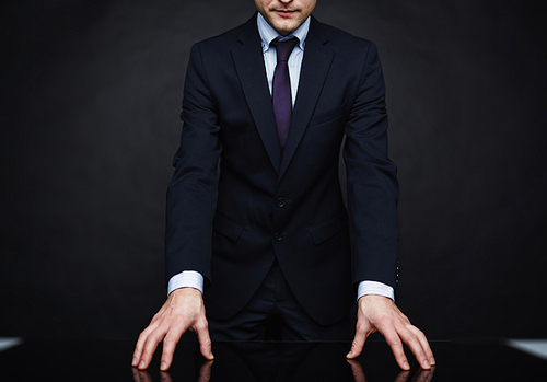 Closeup portrait of unrecognizable authority figure wearing business suit standing leaning on table in powerful pose against black background