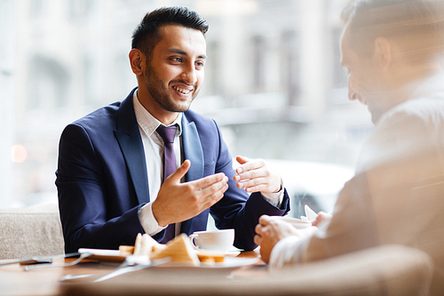 Portrait of enthusiastic young Middle Eastern man gesturing actively and smiling talking to his business partner or colleague in quiet warm lit cafe over cup of coffee