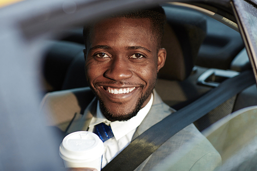 Portrait of confident African -American businessman riding in backseat of car smiling cheerfully  out of window lit by sunlight, holding coffee cup