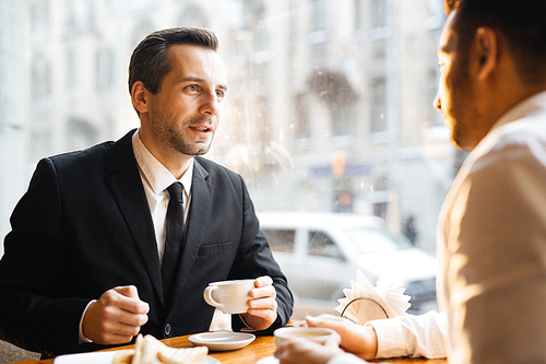 Business meeting at cafe in warm sunset light: two businessmen talking deals over cup of coffee, focus on stylish middle-aged man looking persuasively at his partner