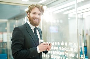 Portrait of young bearded businessman smiling to camera holding smartphone in modern office, copy space